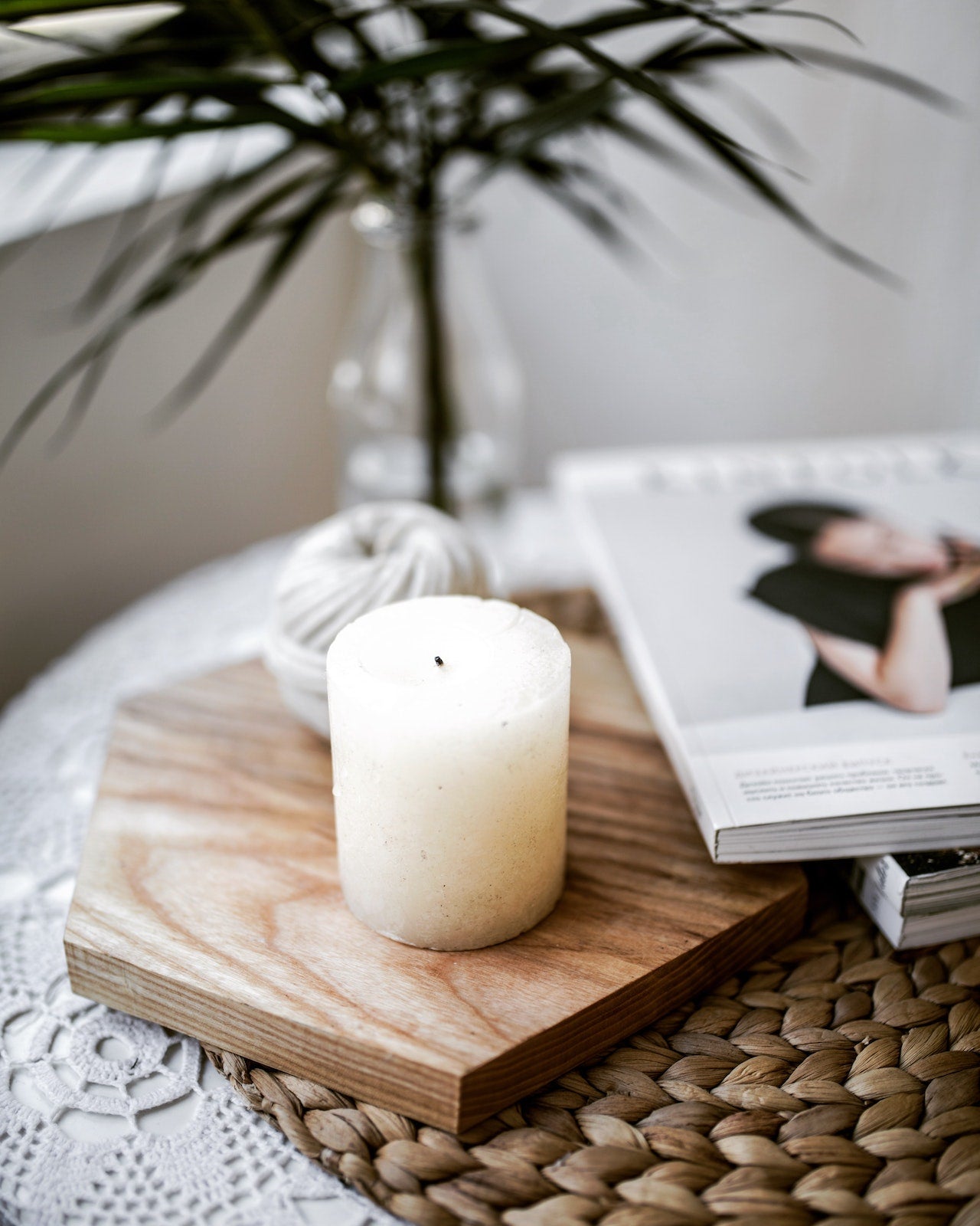 White candle on a wood trivet next to a few magazines. Behind the candle is a vase with a plant.