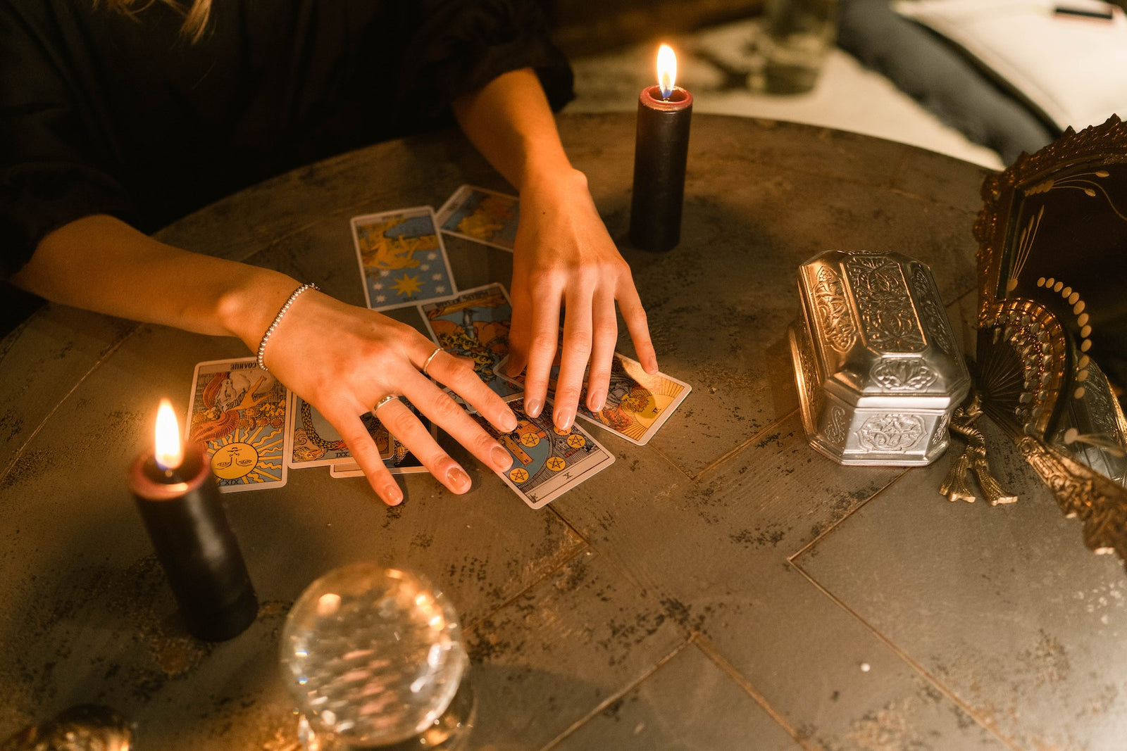 On a table ate to lit black candles, a crystal ball, a small silver box and a persons hands over several face up tarot cards.