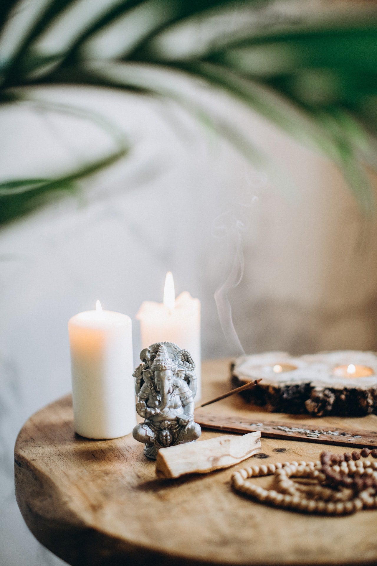 Two lit white candles behind a Ganesha statue with a stick of palo santo in front of it. Next to that is a set of malas and burning incense. 