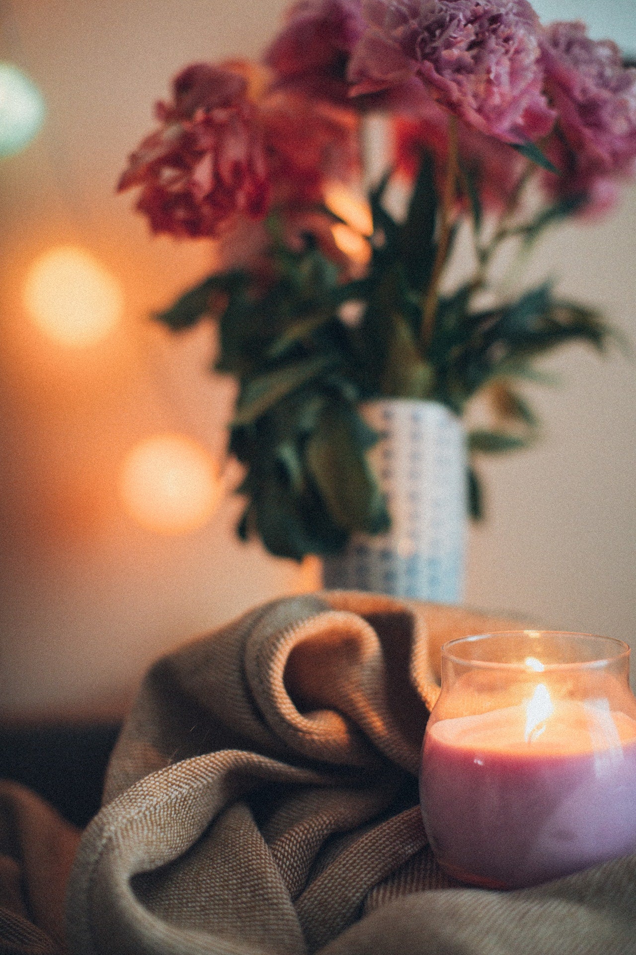 A string of orange-ish white string lights in the background with a vase and purple flowers in the center. In front of that is a cloth and a glass pink lit candle in the front right.