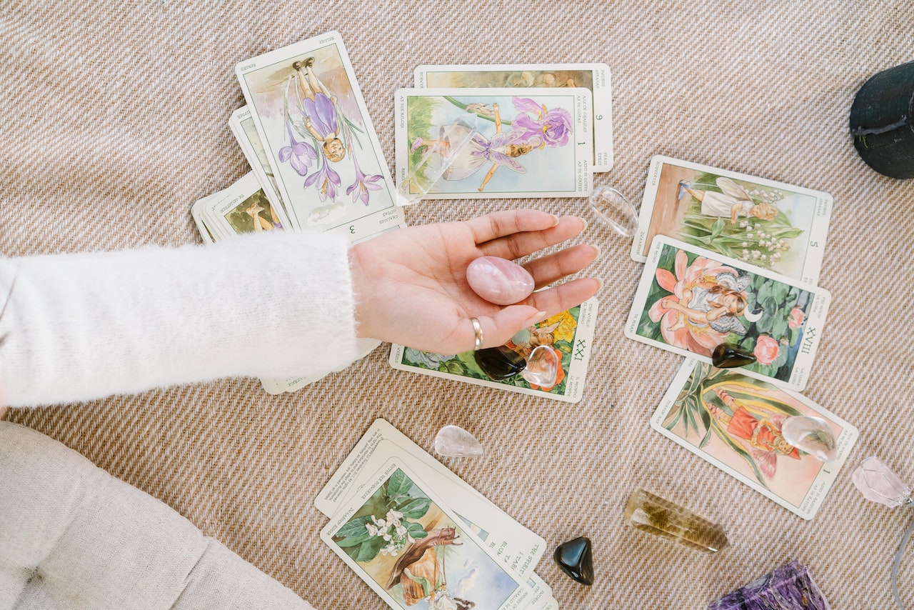 A hand holding a rose quartz crystal over many laid out tarot cards not in any particular order with a few crystals around.