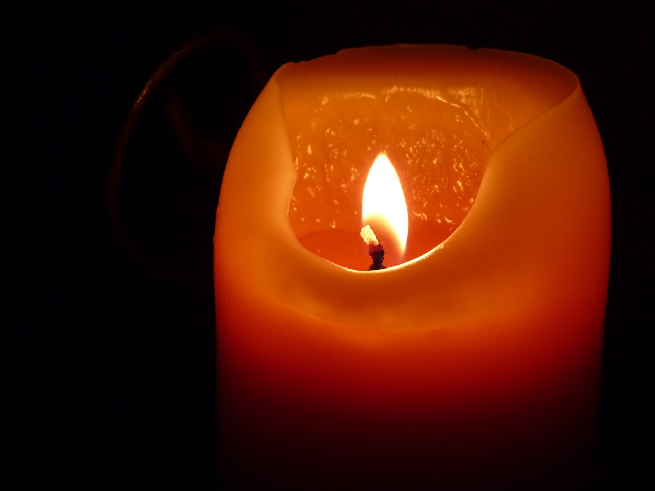 A lit orange pillar candle that has been burning down. A pool of melted wax in the center with one side melted down more then the rest.