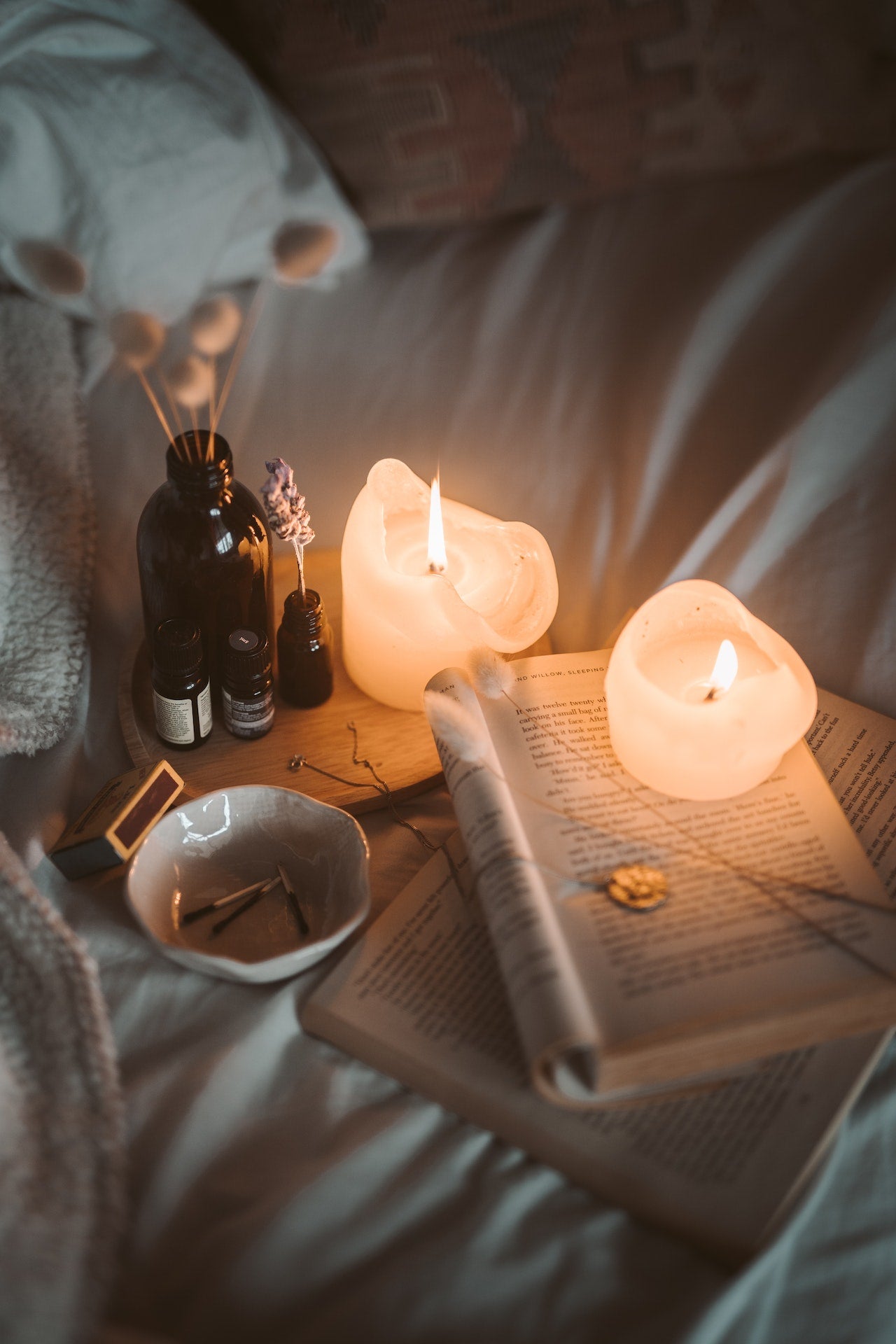 Two lit white candles with 4essential oil’s two of which have flowers in them and an open book. 