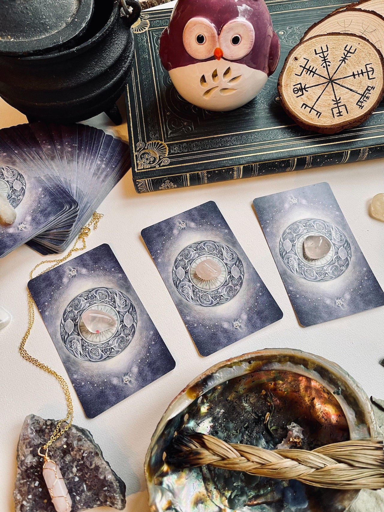 Three tarot cards face down with crystals on each. A cauldron in the top left, a shell and smudge in the bottom right.