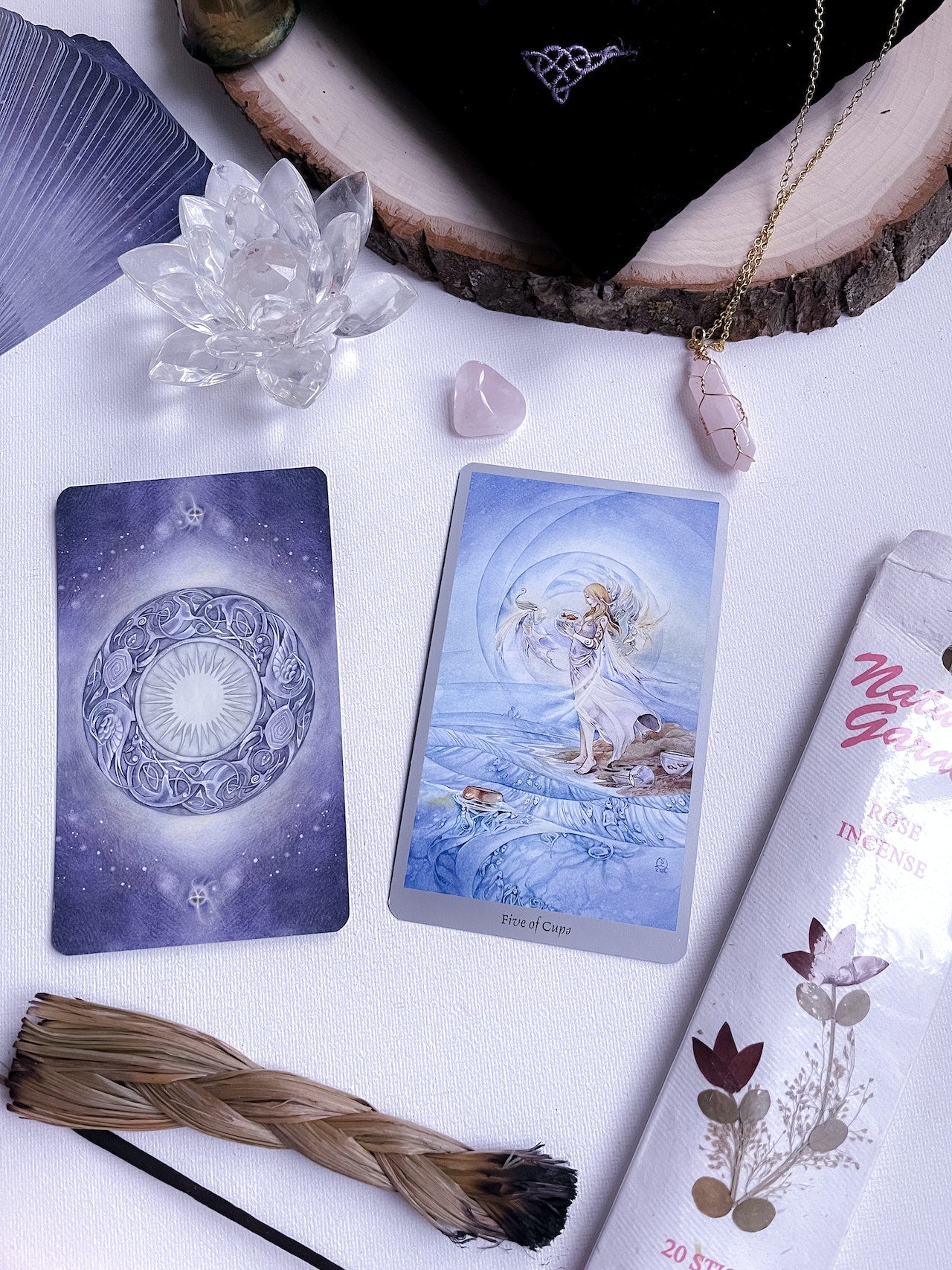 Two tarot cards with one face up and the other face down on a table. The table has sweetgrass and incense laying on the bottom and to the left with crystals around the top above the cards.