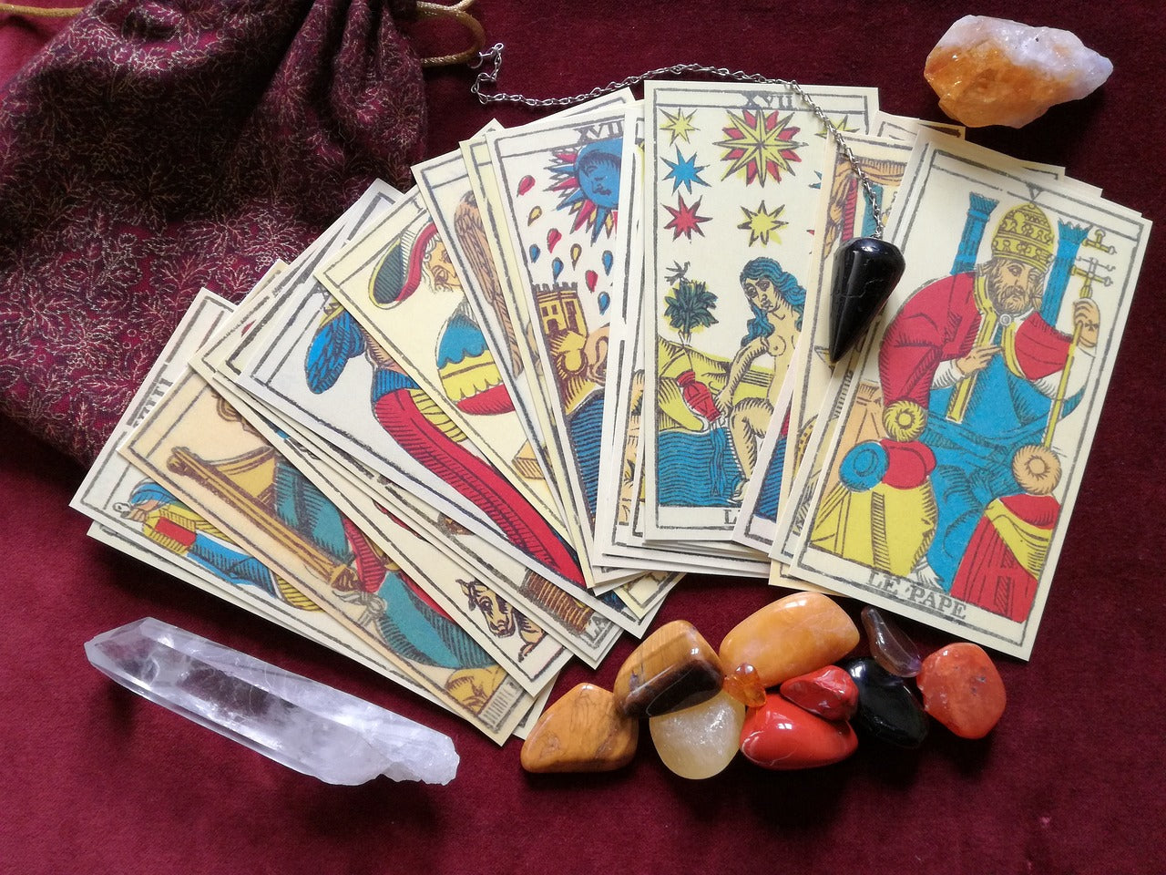 A fanned out row of ten tarot cards with a pendulum and several assorted crystals under the cards and one crystal above the cards on the right side.