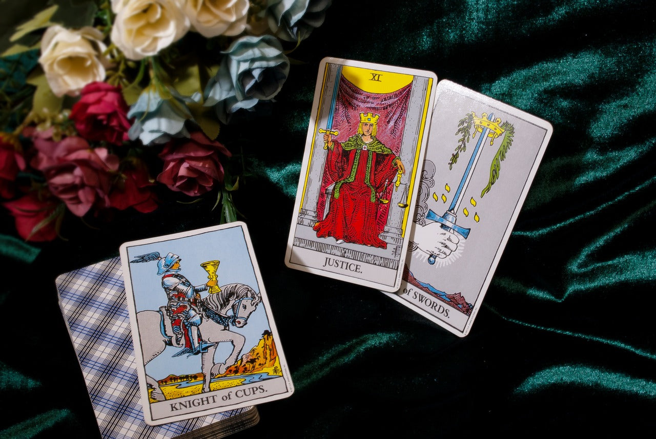 Three tarot cards upright with a stack of cards face down to the left. In the upper left is a gathering of flowers with white, red and green colors. The tarot cards from left to right are the knight of cups, Justice and the ace of swords.