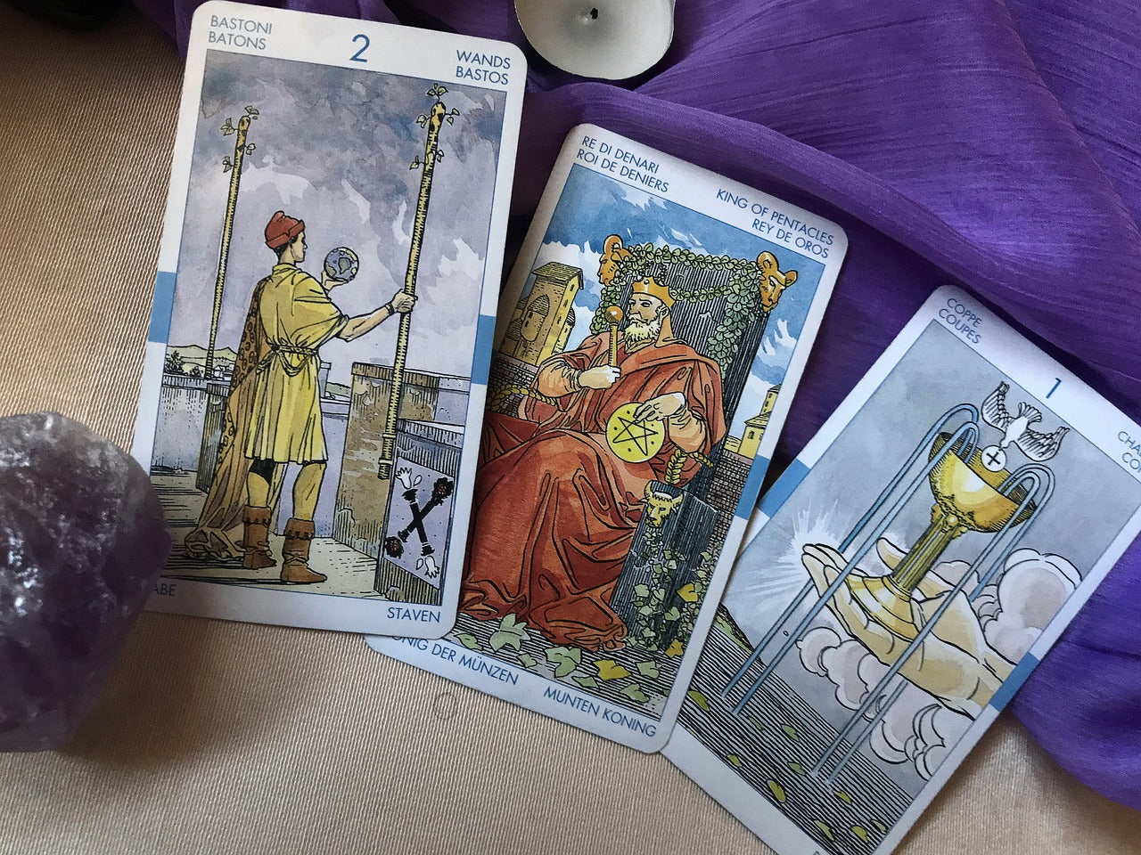 Three tarot cards leaning up against a purple cloth with a piece of amethyst in front of the first card. The three cards are the two of wands, the king of pentacles and the ace of cups.