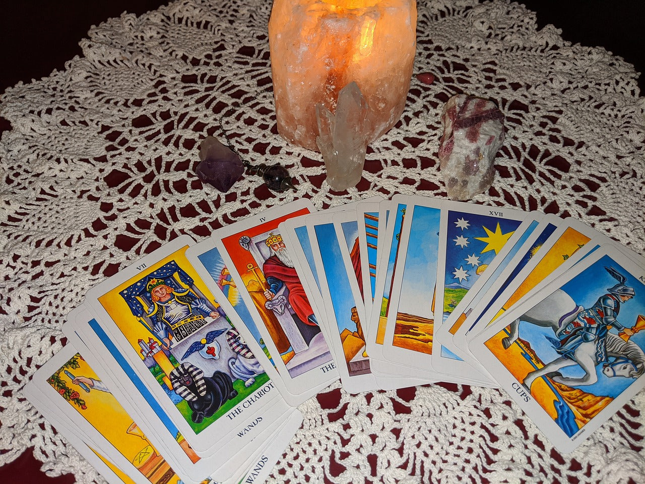 Tarot cards fanned out from left to right on top of a white lace doily. Above the cards on the doily is three different types of crystals and a small lit up salt lamp.