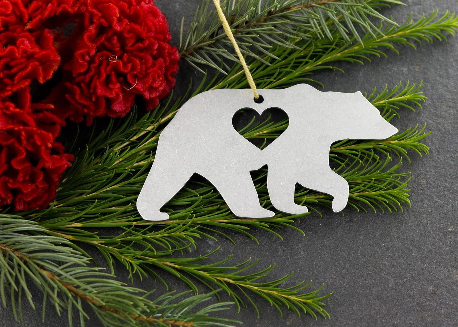 Bear metal ornament with heart cutout, surrounded by greenery and red flowers.