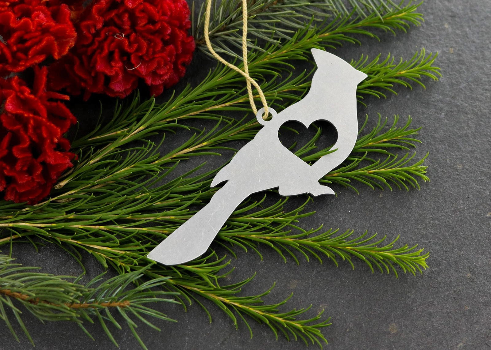 Cardinal metal ornament with heart cutout, surrounded by greenery and red flowers.