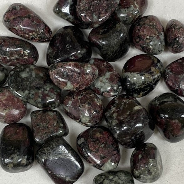 Close-up image of a cluster of tumbled Eudialyte stones with a smooth, polished finish. The stones exhibit a vibrant mix of deep red, black, and white colors, with a speckled and translucent appearance.