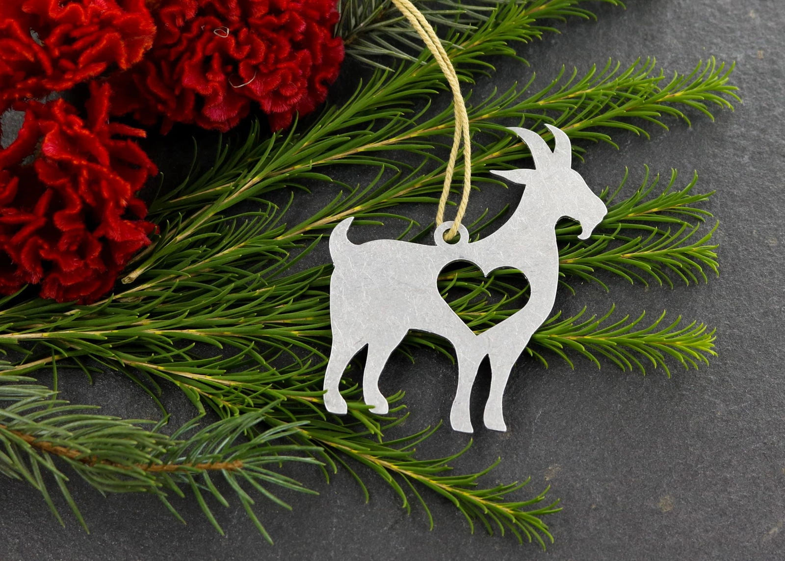 Goat metal ornament with heart cutout, surrounded by greenery and red flowers.