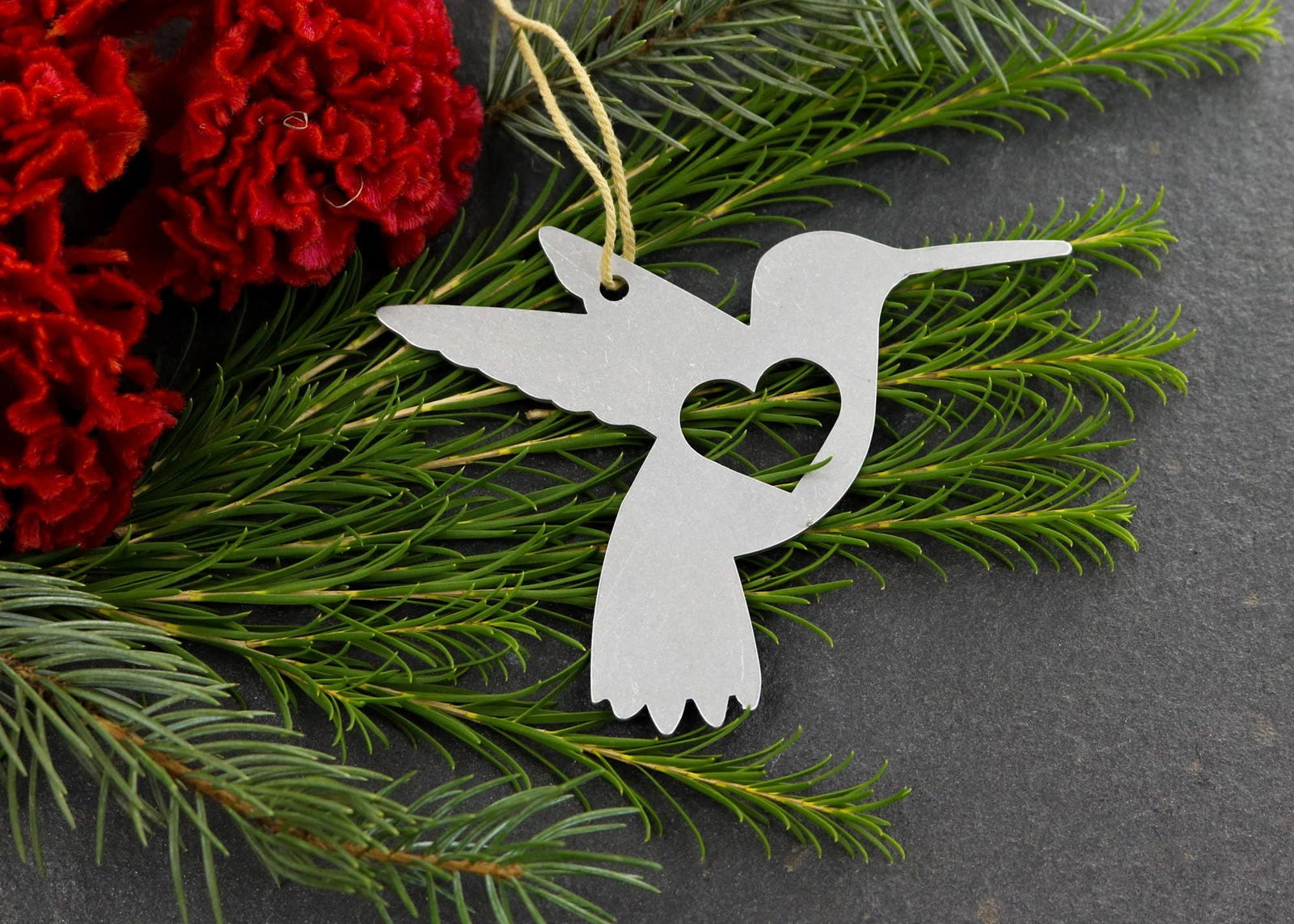 Hummingbird metal ornament with heart cutout, surrounded by greenery and red flowers.