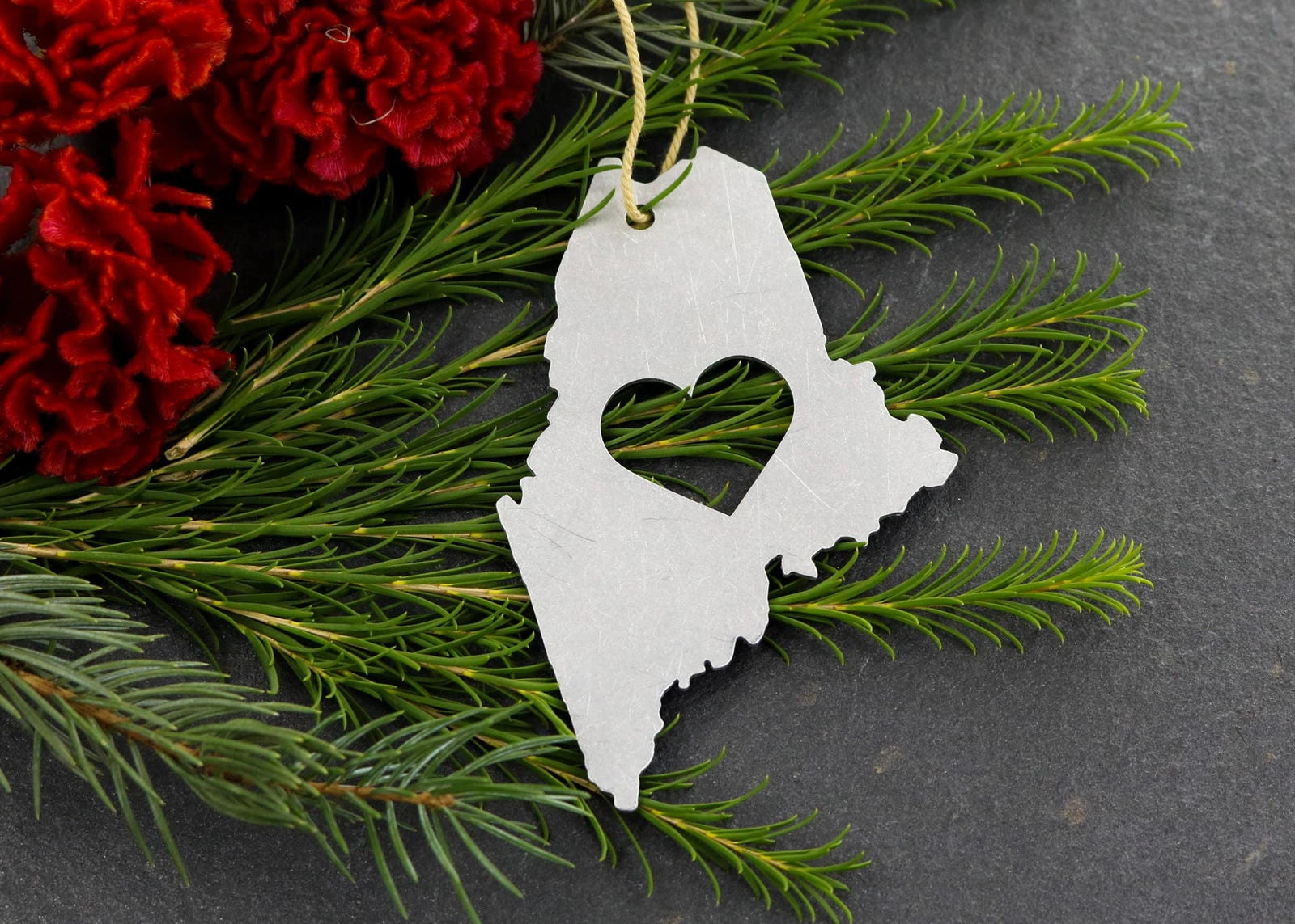 Maine State Metal Ornament with heart cutout, surrounded by greenery.